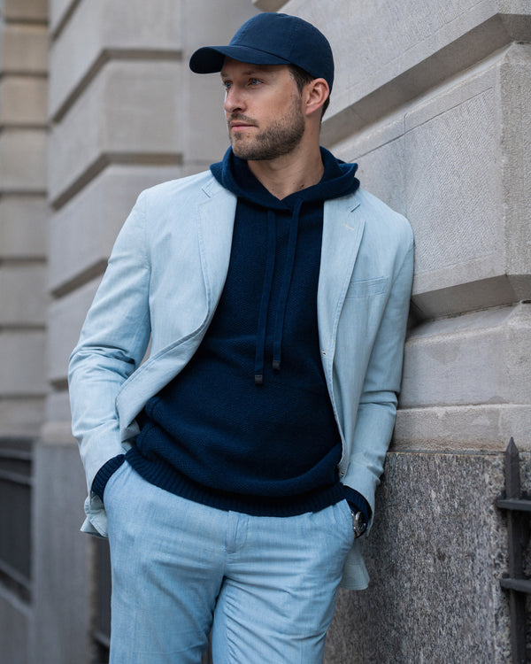 Man leaning against wall wearing blue suit and navy cashmere hoodie and baseball cap