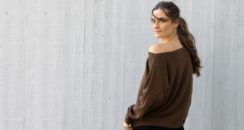 Woman in front of a barn half turned around wearing an off the shoulder brown cashmere sweater