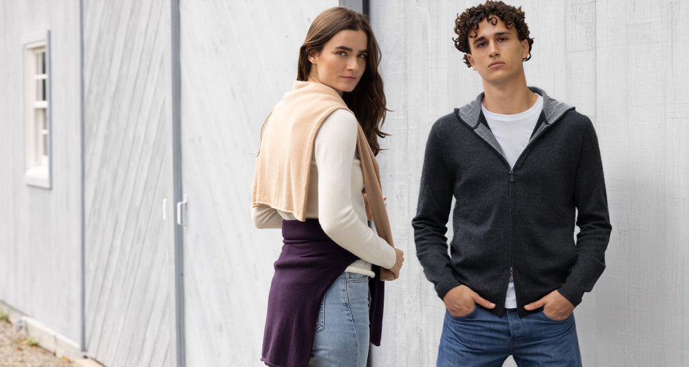 Two people standing outdoors against a light gray wall wearing cashmere sweaters.